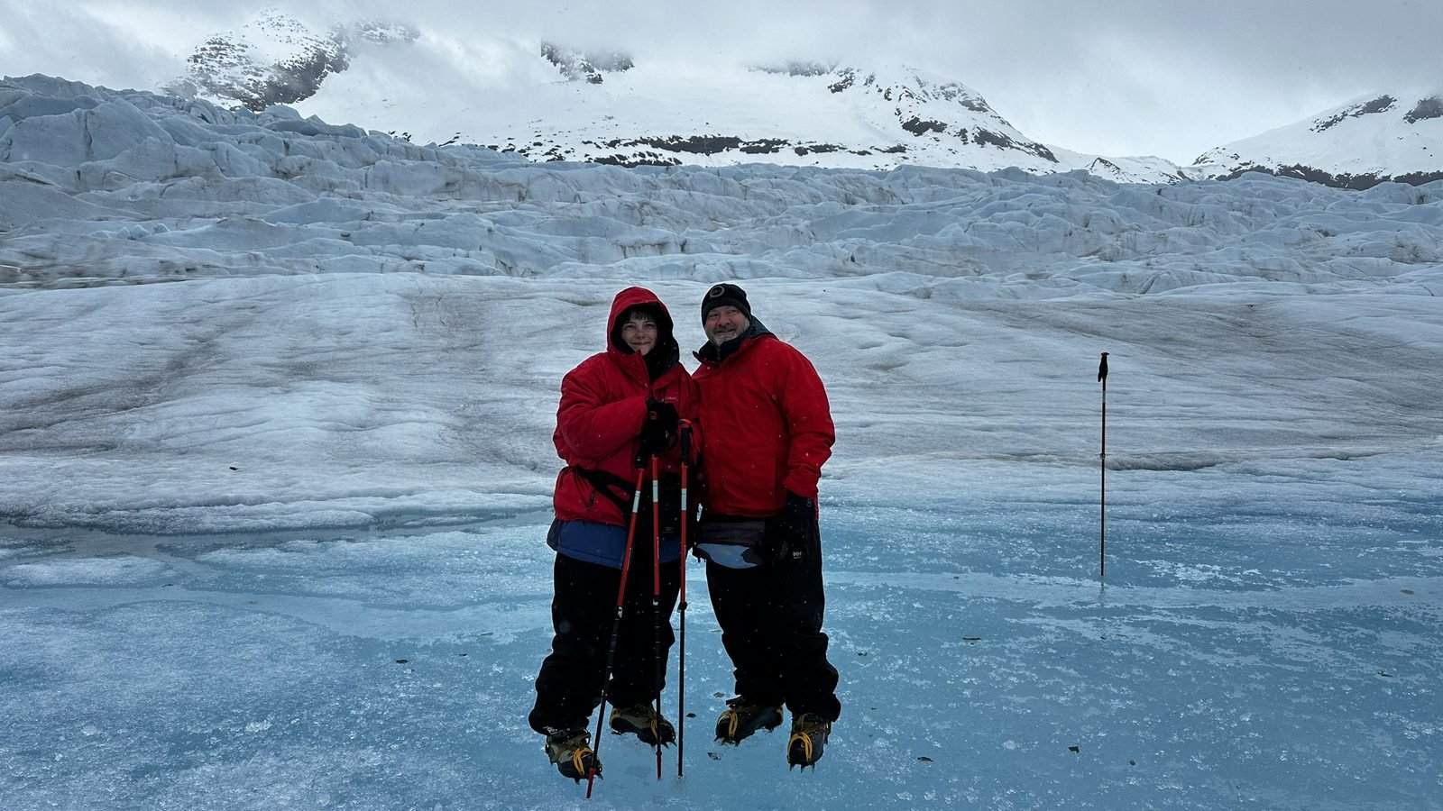 Two hikers in red jackets and crampons standing on blue glacier ice in Alaska, with snow-covered mountains behind them