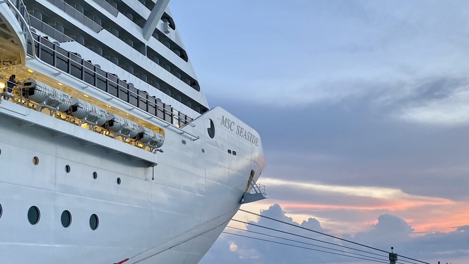 The bow of MSC Seaside at the pier at sunset, with the ship's name on the hull and lit lifeboats along the deck