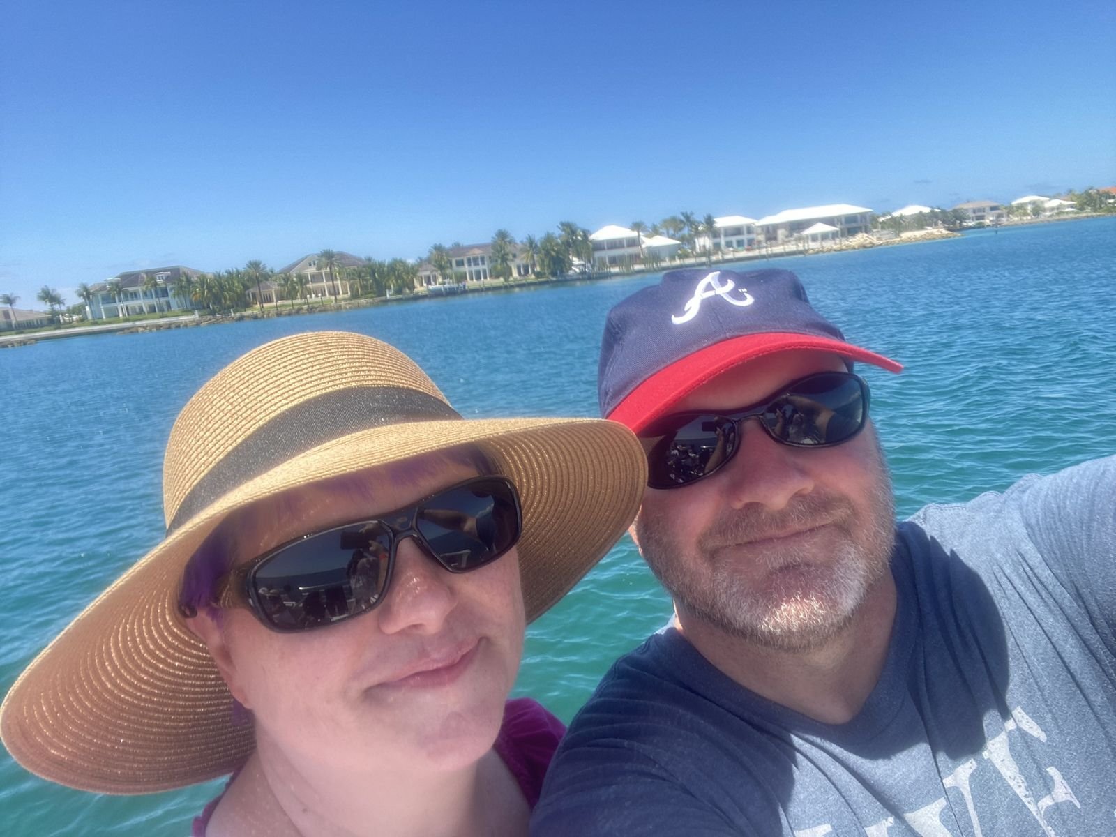 April and Ed Harrell in sunglasses on a boat in turquoise water near Nassau, with palm trees and waterfront homes behind them